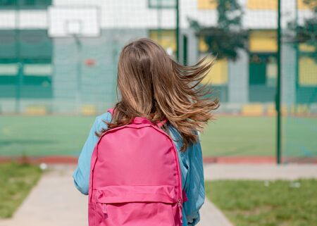 Back view - schoolchild go to school with backpack. Cute child - teen girl with bag against the background of school. Back to school concept.の写真素材