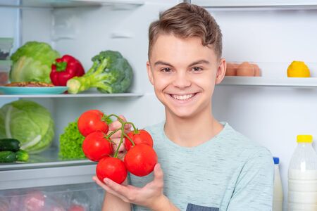 Smiling handsome young teen boy holding fresh red tomatoes while standing near open fridge in kitchen at home. Portrait of pretty child choosing food in refrigerator full of healthy products.の写真素材