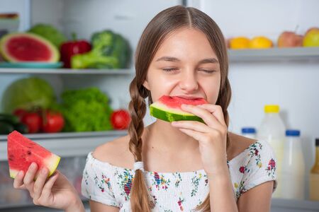 Beautiful young teen girl eating slice ripe red juicy watermelon while standing near open fridge in kitchen at home. Portrait of pretty child choosing food in refrigerator full of healthy products.の写真素材