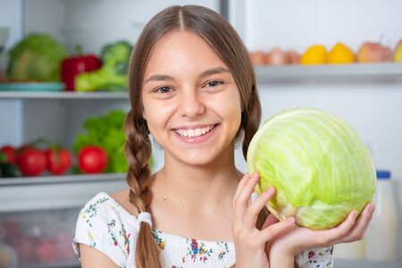 Beautiful young teen girl holding fresh green cabbage while standing near open fridge in kitchen at home. Portrait of pretty smiling child choosing food in refrigerator full of healthy products.の写真素材