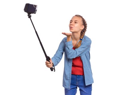 Happy teen girl making selfie with stick and sending an air kiss. Teenager posing to smartphone on selfie stick, isolated on white background. Child taking selfie on mobile phone for social network.の写真素材