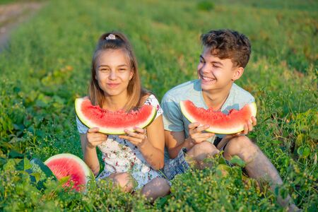 Happy teen boy and girl eating ripe juicy watermelon and smiling. Funny children having picnic in field with slice watermelon. Brother and sister sits on green grass and eat healthy snack in summer.の写真素材