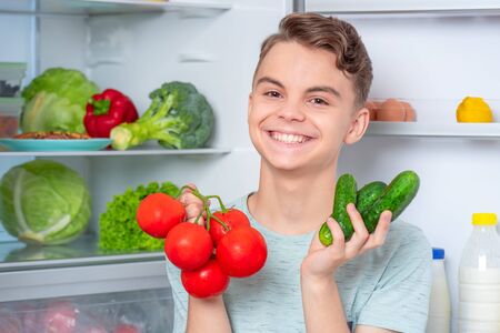 Smiling young teen boy holding fresh red tomatoes and green cucumbers while standing near open fridge in kitchen at home. Portrait of child choosing food in refrigerator full of healthy products.の写真素材