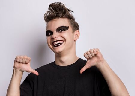 Portrait of happy teen boy with spooking make-up pointing thumbs on himself, on grey background....