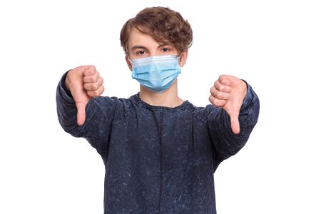 Concept of coronavirus quarantine. COVID-19. Teenager wearing medical protective face mask to health protection from influenza virus, isolated on white background. Teen boy giving thumb down gesture.の写真素材