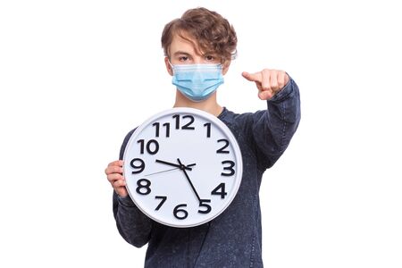 Concept of coronavirus quarantine. Child wearing medical protective face mask during flu virus, holds big clock. COVID-19 - home isolation. Teen Boy pointing fingers at camera, isolated on white.の写真素材