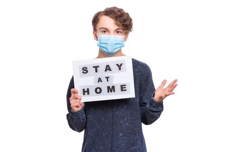 Concept of coronavirus quarantine. Child wearing medical protective face mask during flu virus, holds paper with message for people to stay at home. COVID-19 - self isolation. Boy isolated on white.の写真素材