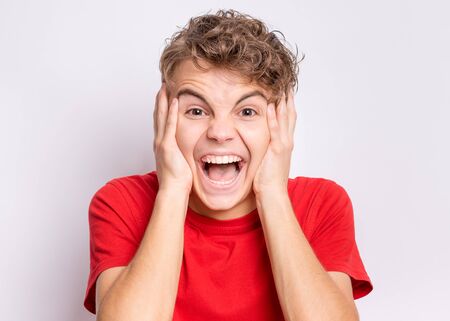 Portrait of surprised teen boy, on gray background. Funny child looking at camera in amazement, touching head with hands. Beautiful caucasian teenager, opening eyes and mouth with shock, screaming.の写真素材