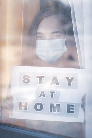 Concept of coronavirus quarantine. Child wearing medical protective face mask during flu virus, looking out of window. COVID-19. Teen girl holds board sign with text Stay at home. View from street.の写真素材