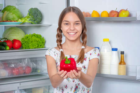 Teen girl holding fresh red pepper while standing near open fridge in kitchen at homeの写真素材