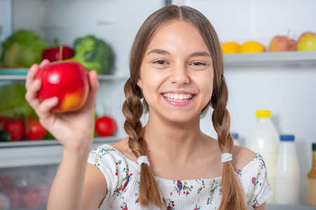 Beautiful young happy teen girl holding fresh red apple while standing near open fridge in kitchen at homeの写真素材
