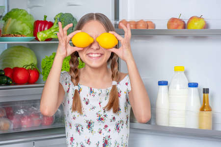Smiling beautiful young teen girl holding yellow lemons over eyes while standing near open fridge in kitchen at homeの写真素材