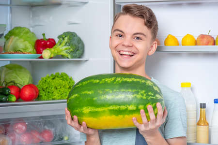 Handsome young teen boy holding big Watermelon while standing near open fridge in kitchen at home. Portrait of pretty child choosing food in refrigerator full of healthy productsの写真素材