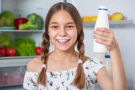 Beautiful young teen girl holding bottle of milk and drinks while standing near open fridge in kitchen at homeの写真素材