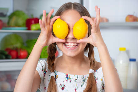 Smiling beautiful young teen girl holding yellow lemons over eyes while standing near open fridge in kitchen at homeの写真素材