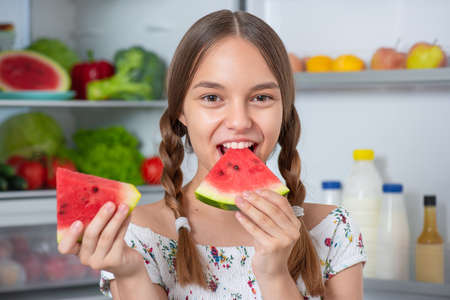 Beautiful young teen girl holding big Watermelon while standing near open fridge in kitchen at homeの写真素材