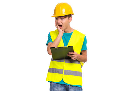 Emotional portrait of teen boy wearing safety jacket and yellow hard hat. Happy cheerful child with notebook and pen pointing something, isolated on white backgroundの写真素材