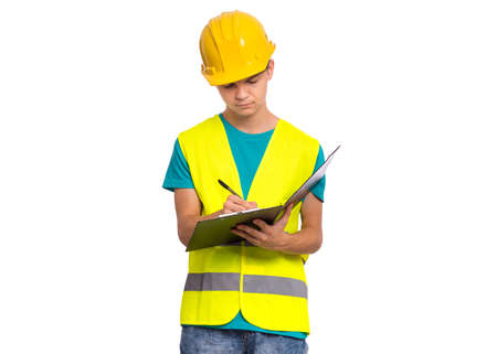 Emotional portrait of teen boy wearing safety jacket and yellow hard hat. Happy cheerful child with notebook and pen pointing something, isolated on white backgroundの写真素材