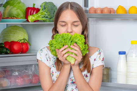 Smiling beautiful young teen girl holding fresh green salad while standing near open fridge in kitchen at homeの写真素材