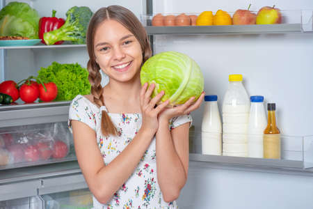 Beautiful young teen girl holding fresh green cabbage while standing near open fridge in kitchen at homeの写真素材