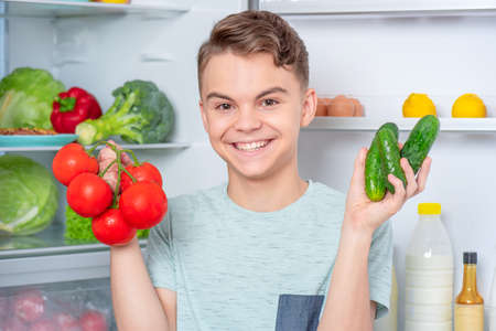 Smiling young teen boy holding fresh red tomatoes and green cucumbers while standing near open fridge in kitchen at home. Portrait of child choosing food in refrigerator full of healthy productsの写真素材