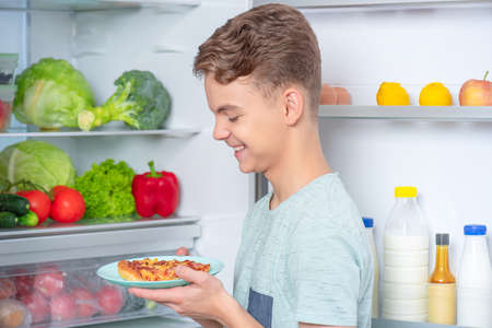 Hungry cute young teen boy holding plate with slice of pizza while standing near open fridge in kitchen at home. Portrait of pretty child choosing food in refrigerator full of healthy productsの写真素材