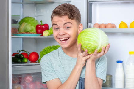 Young teen boy holding fresh green cabbage while standing near open fridge in kitchen at home. Portrait of pretty smiling child choosing food in refrigerator full of healthy productsの写真素材