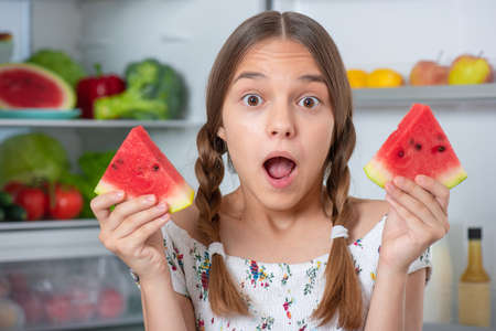 Beautiful young teen girl holding big Watermelon while standing near open fridge in kitchen at homeの写真素材