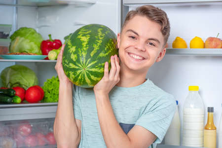 Handsome young teen boy holding big Watermelon while standing near open fridge in kitchen at home. Portrait of pretty child choosing food in refrigerator full of healthy productsの写真素材