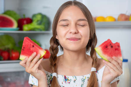 Beautiful young teen girl holding big Watermelon while standing near open fridge in kitchen at homeの写真素材