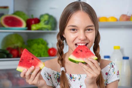 Beautiful young teen girl holding big Watermelon while standing near open fridge in kitchen at homeの写真素材