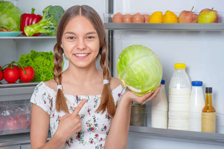 Beautiful young teen girl holding fresh green cabbage while standing near open fridge in kitchen at homeの写真素材