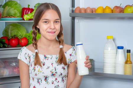 Beautiful young teen girl holding bottle of milk and drinks while standing near open fridge in kitchen at homeの写真素材