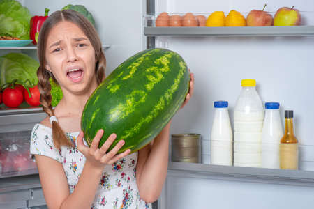 Beautiful young teen girl holding big Watermelon while standing near open fridge in kitchen at homeの写真素材
