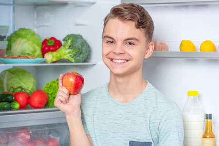 Smiling handsome young teen boy holding fresh red apple while standing near open fridge in kitchen at home. Portrait of pretty child choosing food in refrigerator full of healthy productsの写真素材