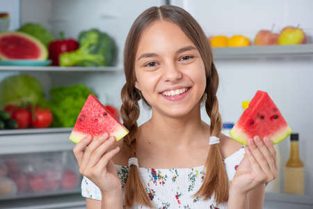 Beautiful young teen girl holding big Watermelon while standing near open fridge in kitchen at homeの写真素材