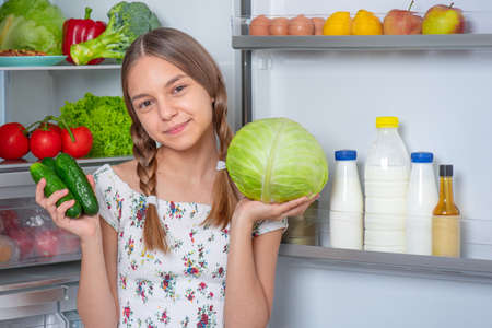 Beautiful young teen girl holding fresh green cabbage and green cucumbers while standing near open fridge in kitchen at homeの写真素材