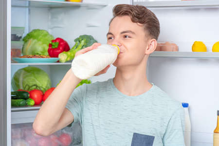 Cute young teen boy holding bottle of milk and drinks while standing near open fridge in kitchen at home. Portrait of pretty child choosing food in refrigerator full of healthy productsの写真素材