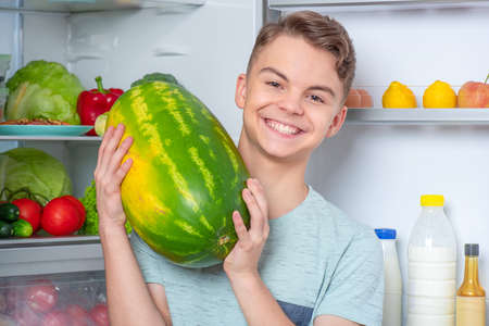Handsome young teen boy holding big Watermelon while standing near open fridge in kitchen at home. Portrait of pretty child choosing food in refrigerator full of healthy productsの写真素材