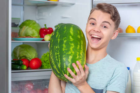 Handsome young teen boy holding big Watermelon while standing near open fridge in kitchen at home. Portrait of pretty child choosing food in refrigerator full of healthy productsの写真素材
