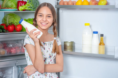 Beautiful young teen girl holding bottle of milk and drinks while standing near open fridge in kitchen at homeの写真素材
