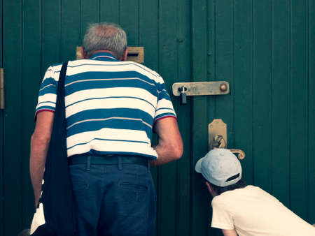 Senior and child are looking through the mail slot and keyhole, view from the back. Grandfather and grandson see something interesting. Concept of two generations, peeping and peekingの写真素材