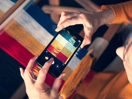 Artisan holding device and making picture of her weaving product for social networks. Weaver taking photo with smartphone, selective focusの写真素材