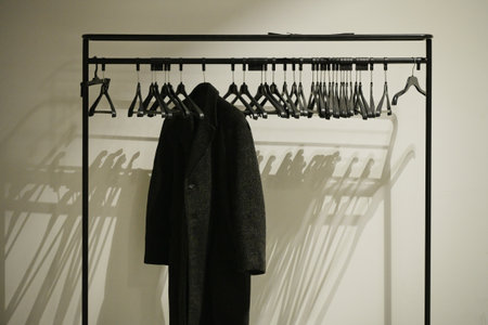 A long black coat hangs on a hanger on a rack, against a white wall.の写真素材