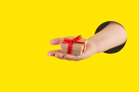 Hand holds a gift box made of natural materials through a hole in a yellow paper background. Template for design. Close up and horizontal orientation. Copy space.の写真素材