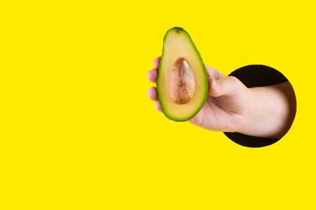 Hand holds half cut avocado through a hole in a yellow paper background. Template for design. Close up and horizontal orientation. Copy space. の写真素材