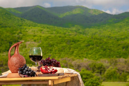 Outdoor picnics in the mountains. A picnic table set with red wine, cheese, fruits, grapes and bread stands in a meadow in green grass. The concept of secluded outdoor recreation.の写真素材