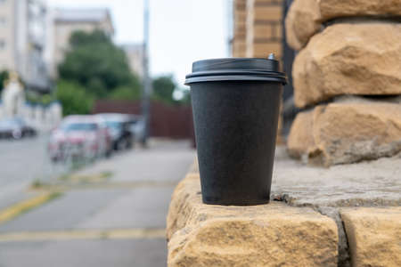 A black paper cup with coffee stands on a brick wall outside. Concepts for a coffee shop.