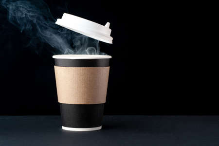 Black cup of coffee with a white cap on a black background. Hot drink with steam. Recycled and...