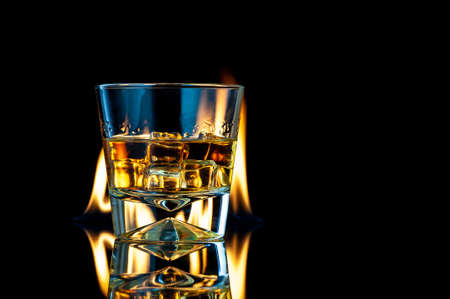 Whiskey or bourbon in a transparent glass with ice cubes on a black background with fire. Horizontal template for design. Place for your text.の写真素材